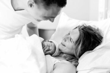 Vrouw is thuis bevallen en heeft pasgeboren baby op borst en partner kijkt liefdevol