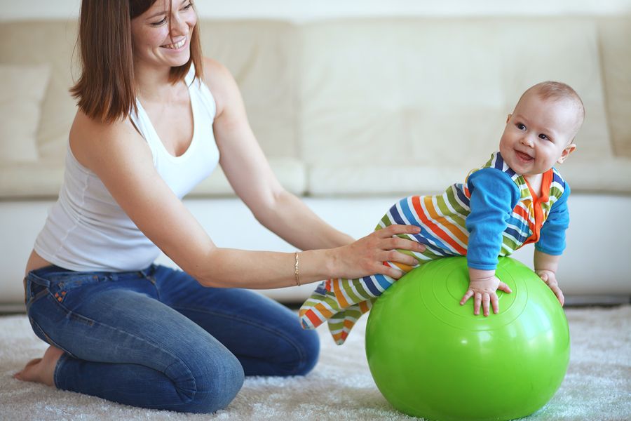 Lachende baby van 26 weken oud ligt op fitness bal, moeder houdt hem bij zijn heupjes vast