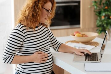 Zwangere vrouw staat met laptop aan keukentafel om dingen te regelen voor haar zwangerschap en bevalling