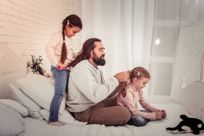 Vader zit met dochters op bed, hij vlecht haar jongste dochter en zijn haar wordt gevlochten door oudste dochter