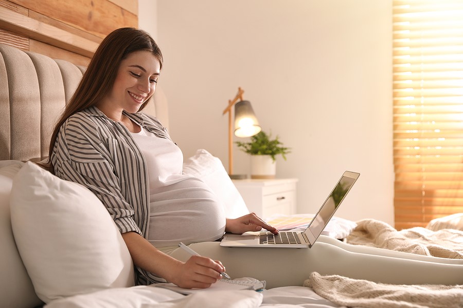 Schwangere liegt mit Computer und Notizbuch im Bett und macht sich Notizen